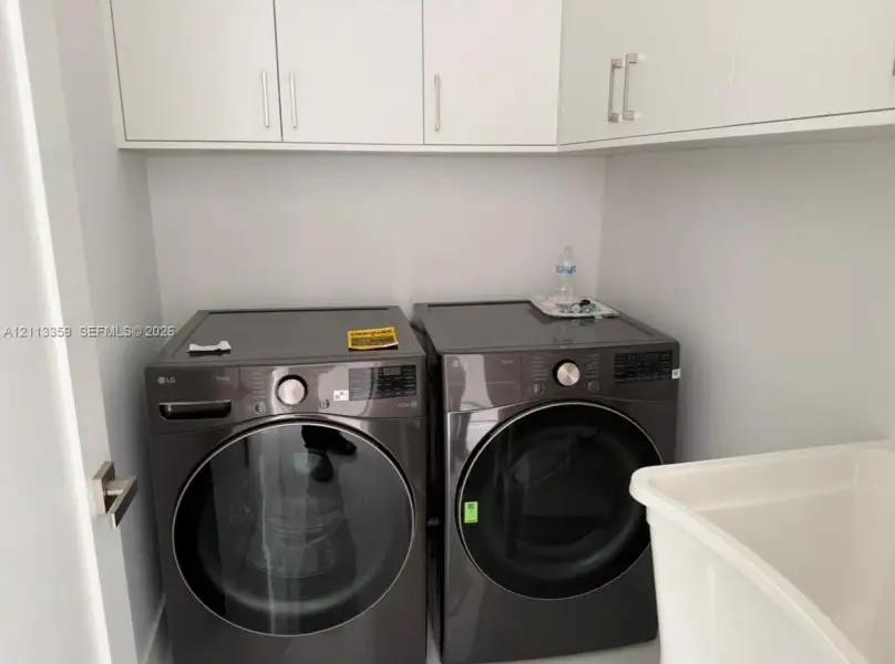 Laundry space featuring charcoal-finish front-load appliances, matte white shaker-style cabinetry with polished metal hardware, and a deep basin utility sink