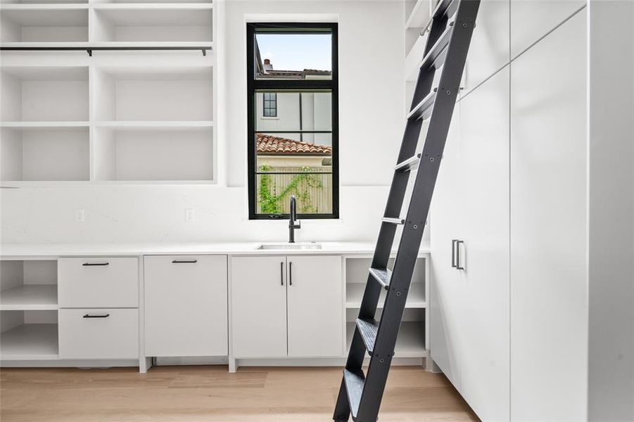 The scullery is a dedicated space for food storage and preparation. Bathed in natural light from the windows, this functional haven includes a sink, dishwasher, compact refrigerator, built-in shelving, and a sliding ladder for easy access to higher shelves.