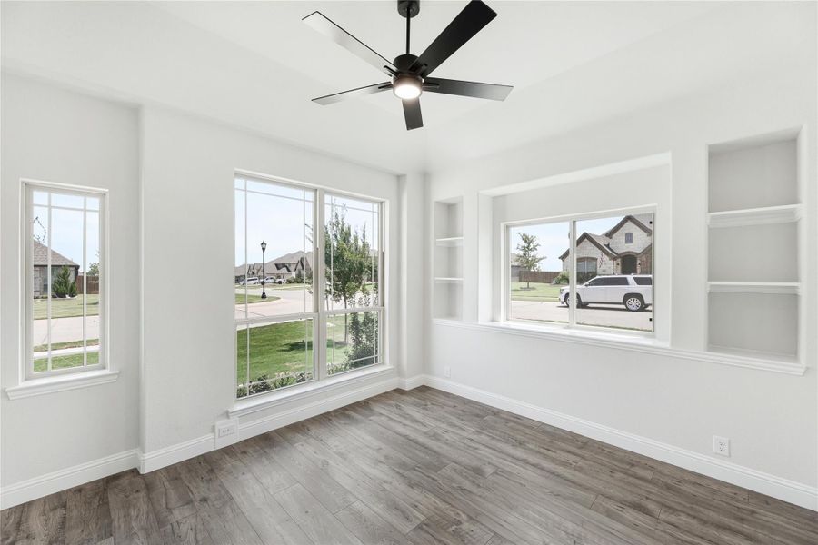 Empty room with hardwood floors, ceiling fan, built-in shelves, and large windows overlooking a neighborhood