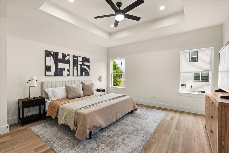 Primary bedroom with tray ceiling, modern fan, and tall windows overlooking greenery Primary bedroom with tray ceiling, modern fan, and tall windows overlooking greenery