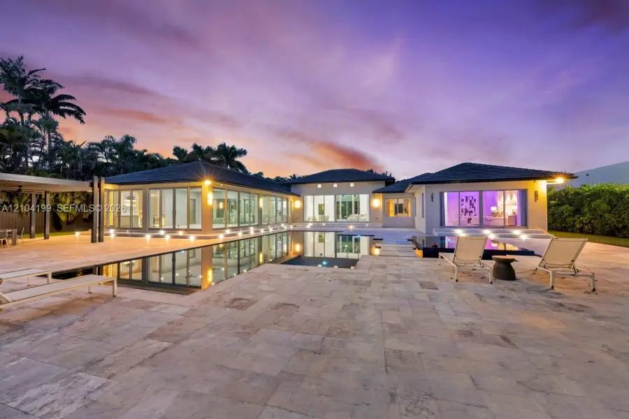 Single-story residence featuring an expansive stone-paved patio, an infinity-edge swimming pool, extensive floor-to-ceiling glass wall panels, and recessed exterior lighting