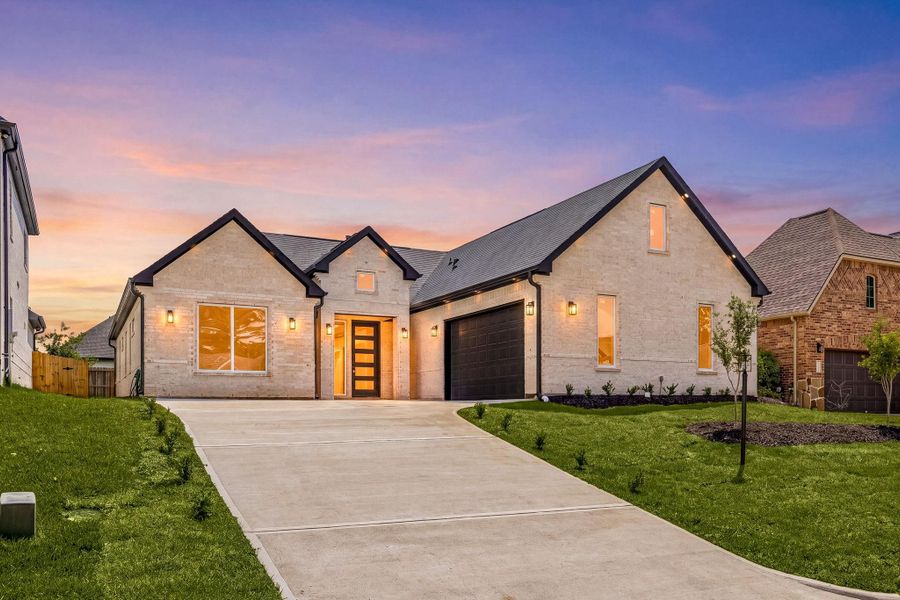 This photo showcases a modern single-story home with a clean, light brick facade and dark roof accents. It features a spacious driveway leading to a two-car garage and a well-maintained front lawn. The house is warmly lit, enhancing its curb appeal against a colorful sunset sky.