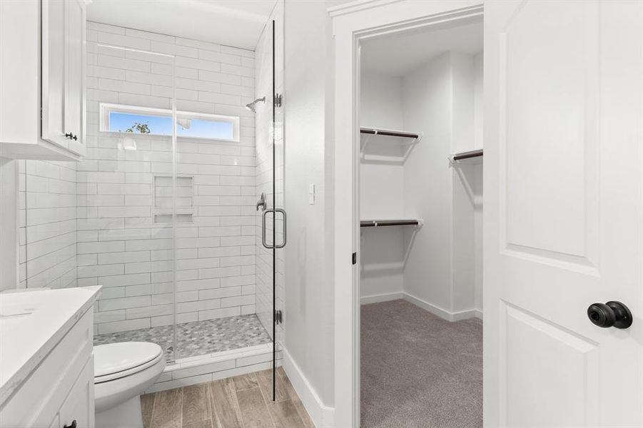 This is a modern bathroom with a walk-in shower featuring subway tiles and a pebble floor. There is a white vanity and toilet, with a space-saving sliding glass shower door. A small window allows for natural light. Adjacent to the bathroom is a walk-in closet with built-in shelves for storage.