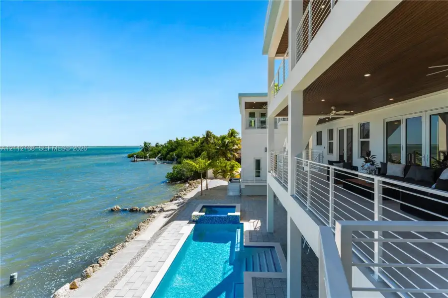 Multi-level waterfront residence featuring a lap pool with an integrated spa, expansive covered balcony with slatted wood-finish ceilings, metal railings, and stone-paver hardscaping Multi-level waterfront residence featuring a lap pool with an integrated spa, expansive covered balcony with slatted wood-finish ceilings, metal railings, and stone-paver hardscaping