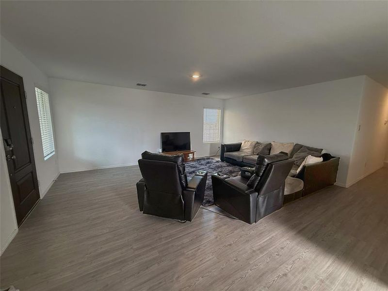 Spacious living area featuring light wood-finish flooring, white walls, and a dark paneled entry door Spacious living area featuring light wood-finish flooring, white walls, and a dark paneled entry door