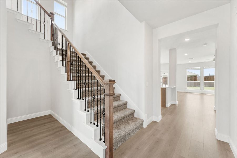 Carpeted staircase with wood handrail and iron balusters in an open entryway with light hardwood floors