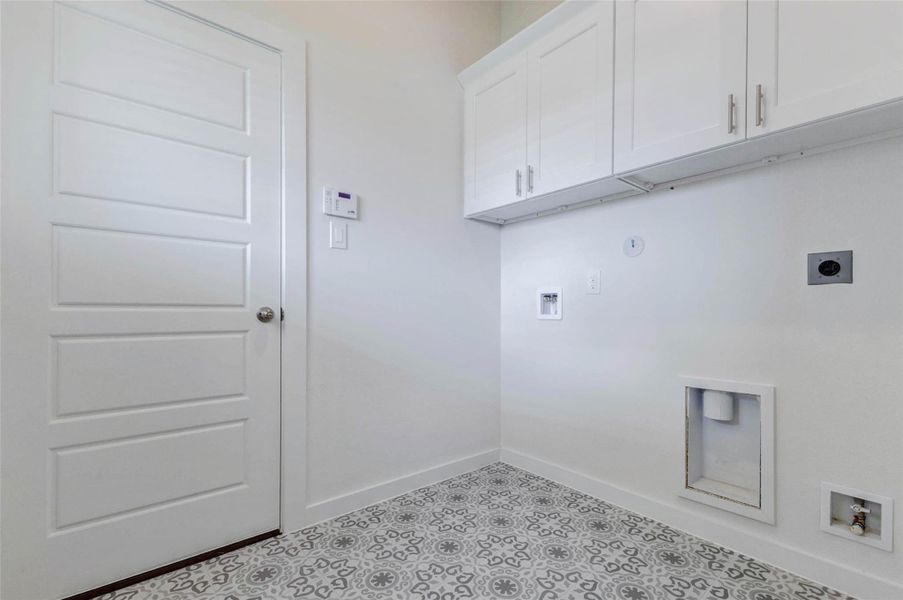 Utility room with patterned tile flooring, washer and dryer hookups, upper storage cabinets, exterior-style door, and wall-mounted thermostat nearby. This dedicated laundry space offers practical function with room for appliances, cleaning supplies, and household storage while keeping laundry conveniently located inside the home.