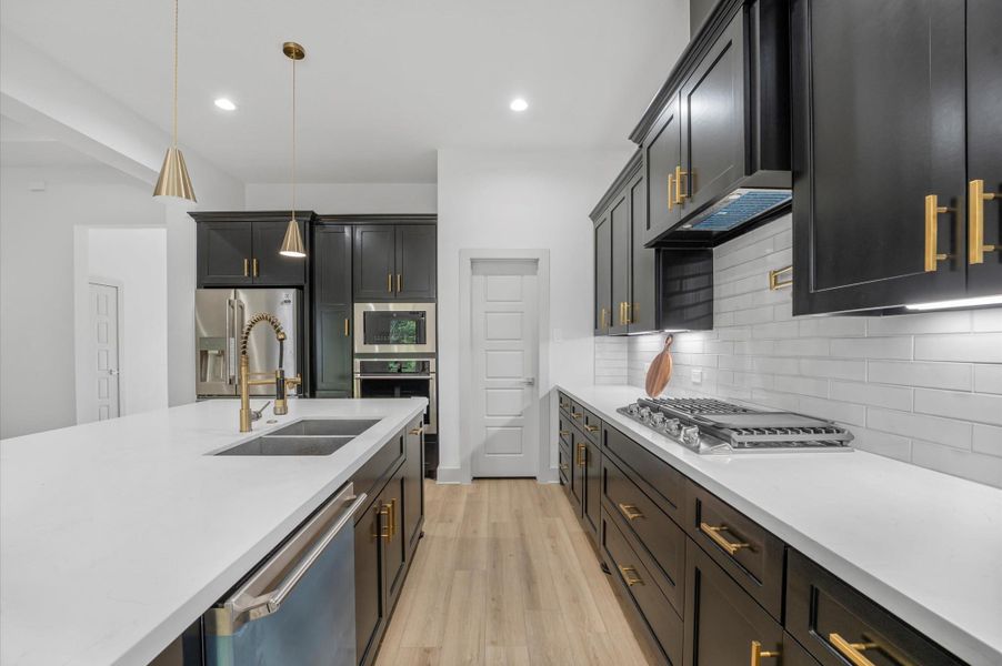 This kitchen features sleek black cabinetry with gold handles, a modern white countertop, and a stylish backsplash. It includes a built-in oven, gas stovetop, stainless steel fridge, and a dishwasher. The space is brightened by pendant lighting and an open layout. This kitchen features sleek black cabinetry with gold handles, a modern white countertop, and a stylish backsplash. It includes a built-in oven, gas stovetop, stainless steel fridge, and a dishwasher. The space is brightened by pendant lighting and an open layout.