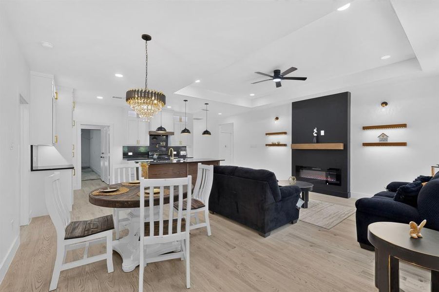 Open-concept living space featuring wood-finish flooring throughout, a modern fireplace with a dark accent wall and wood mantel, and tray ceilings