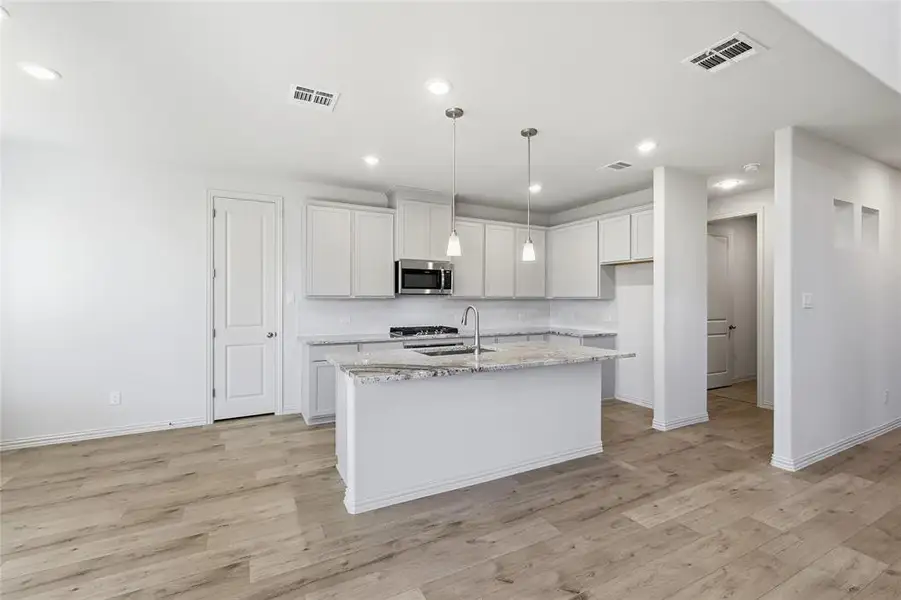 Kitchen featuring decorative light fixtures, a center island with sink, stainless steel microwave, light stone countertops, and white cabinets