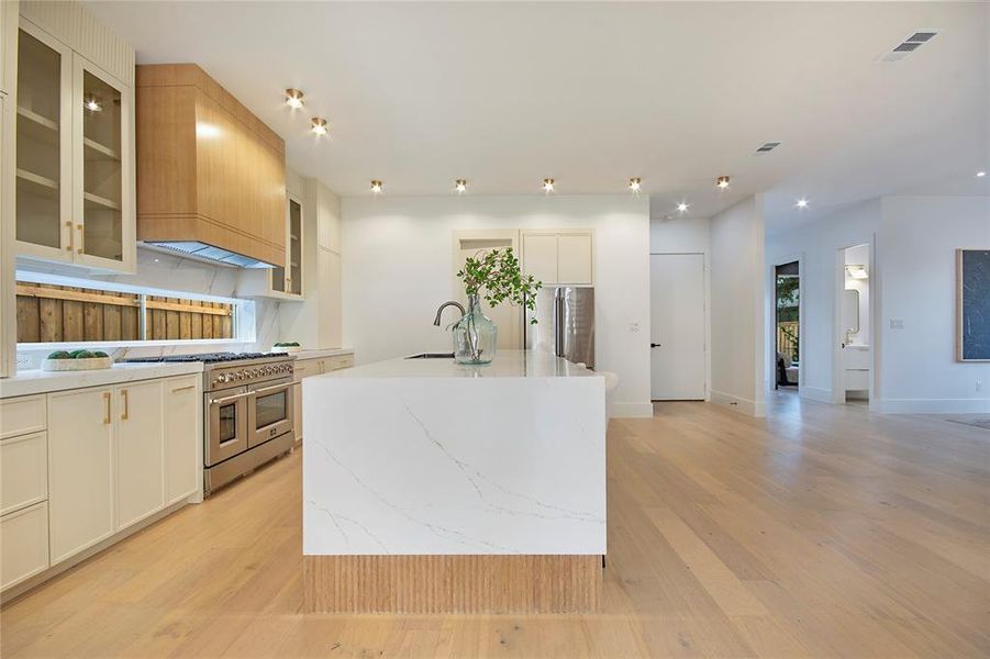 Kitchen with a kitchen island with sink, light wood-style floors, appliances with stainless steel finishes, premium range hood, and light stone counters Kitchen with a kitchen island with sink, light wood-style floors, appliances with stainless steel finishes, premium range hood, and light stone counters