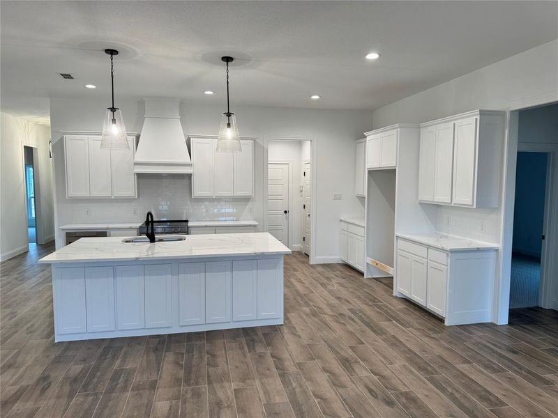 Kitchen featuring white cabinetry, decorative light fixtures, an island with sink, and custom range hood