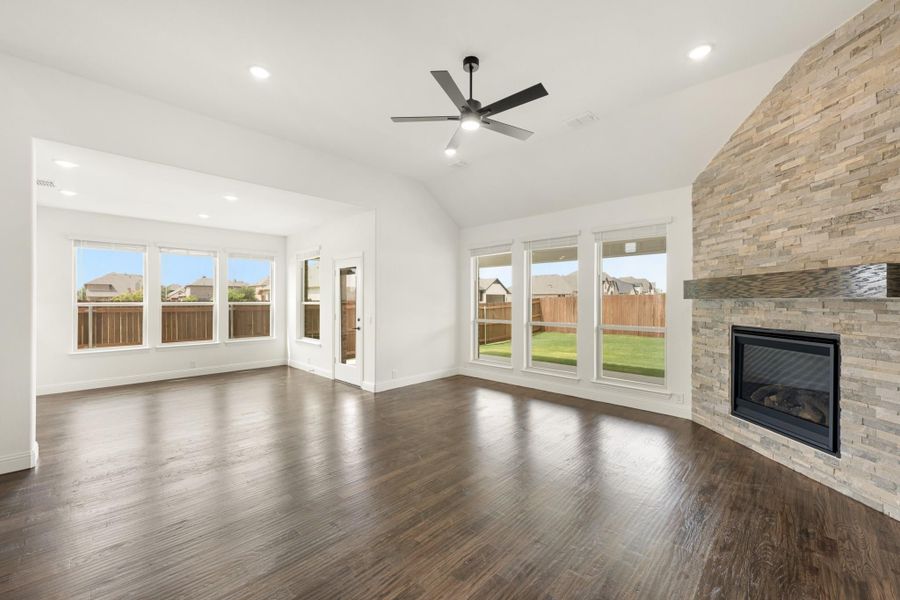 Open living room with stone fireplace, dark hardwood floors, ceiling fan, and large backyard-facing windows