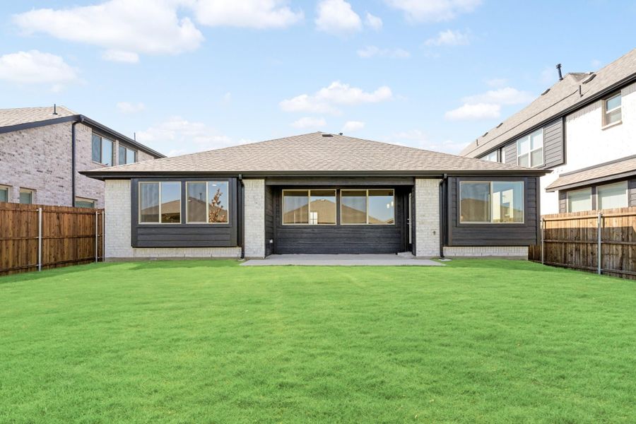 Rear exterior of single-story home with dark siding, large windows, patio, and fenced green backyard