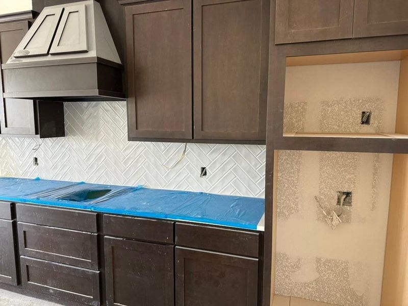Kitchen featuring dark wood-finish cabinetry, a light herringbone tile backsplash, and a built-in range hood with light gray accents