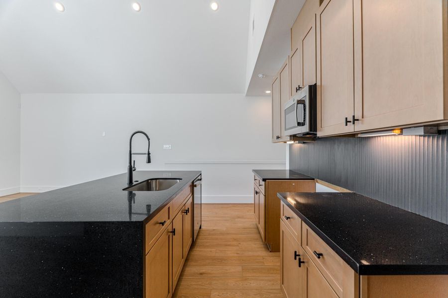 This kitchen features modern wood cabinets, sleek black countertops, and a central island with a sink and high-arc faucet. It has a built-in microwave and ambient lighting, creating a stylish, functional space.Photo Disclaimer: Photos are representative of the community and may depict a similar floor plan, model home, or previously completed residence. This kitchen features modern wood cabinets, sleek black countertops, and a central island with a sink and high-arc faucet. It has a built-in microwave and ambient lighting, creating a stylish, functional space.Photo Disclaimer: Photos are representative of the community and may depict a similar floor plan, model home, or previously completed residence.