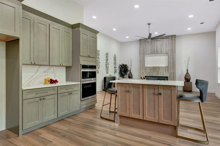 Open-concept interior featuring wood-finish flooring, a kitchen with two-tone cabinetry and built-in double ovens, a central island with a light-toned countertop, and a fireplace with a textured wood-look surround Open-concept interior featuring wood-finish flooring, a kitchen with two-tone cabinetry and built-in double ovens, a central island with a light-toned countertop, and a fireplace with a textured wood-look surround
