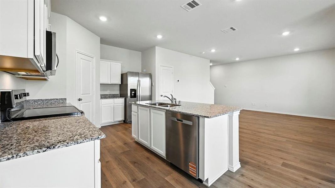 Open-concept kitchen featuring an island with integrated sink, stainless steel appliances, white cabinetry, granite countertops, and wood-finish flooring