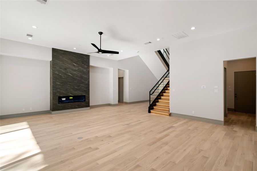 Spacious modern living area with light wood flooring, a sleek black fireplace, recessed lighting, and a striking staircase with illuminated steps. Perfect for contemporary living.