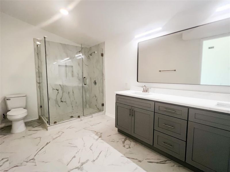 Spacious bathroom featuring a frameless glass shower enclosure with marbled wall tile, a double vanity with grey cabinetry and white countertops, a large rectangular mirror, and marbled floor tiling