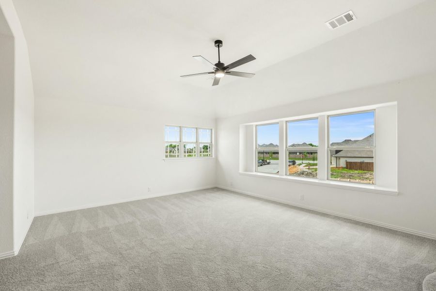 Empty room with vaulted ceiling, ceiling fan, gray carpet, and large windows with neighborhood view Empty room with vaulted ceiling, ceiling fan, gray carpet, and large windows with neighborhood view