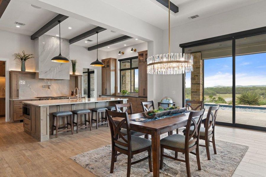 Open-concept kitchen and dining area featuring wood-finish flooring, a large kitchen island with seating, a patterned stone backsplash and range hood, exposed ceiling beams, and a dining area with a contemporary chandelier