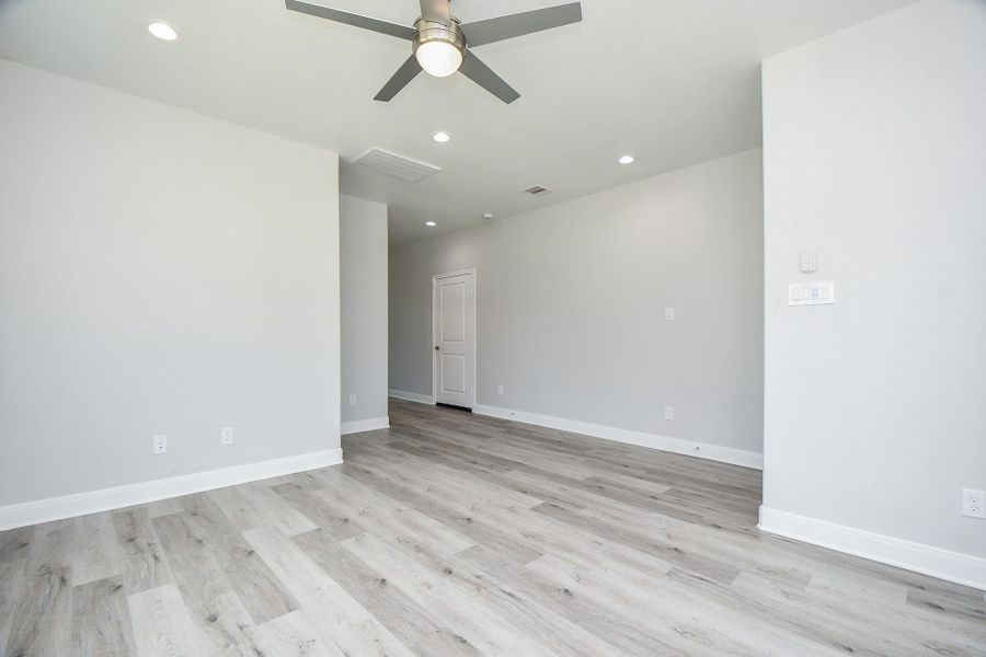 This is a spacious, main living room featuring light gray walls, contemporary wood-look flooring, and a ceiling fan with lighting. There's ample natural light and a doorway leading to another part of the home. The room is a blank canvas ready for personalization.