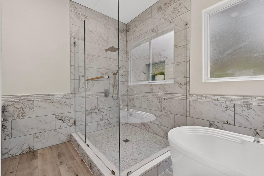 Floor-to-ceiling marble-look wall tile encloses a glass-walled shower beside a freestanding soaking tub.
