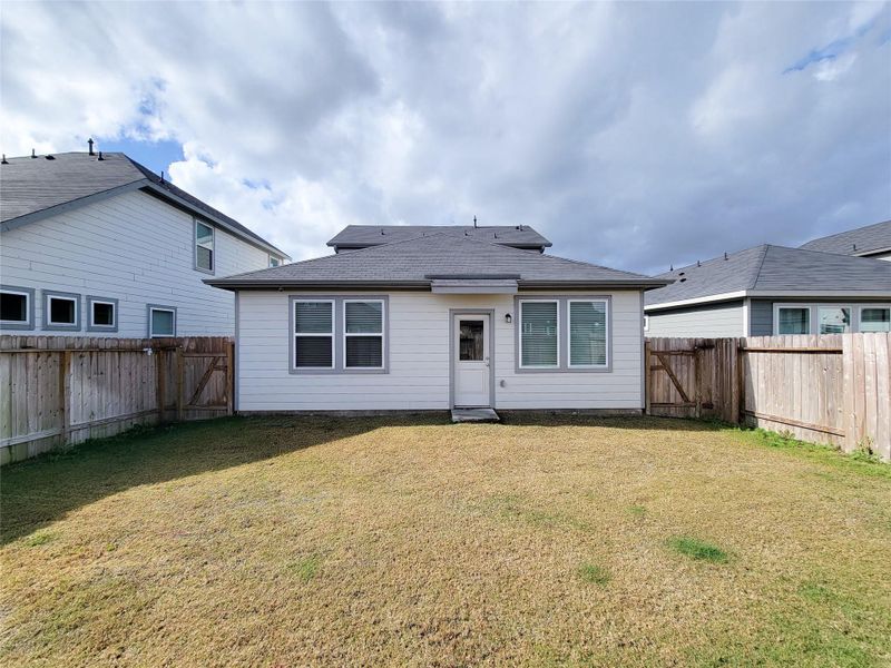 White siding and a dark roof. It has a small fenced backyard with a grassy area, a central back door, and two large windows. The space is enclosed by wooden fencing, providing privacy from neighboring homes.