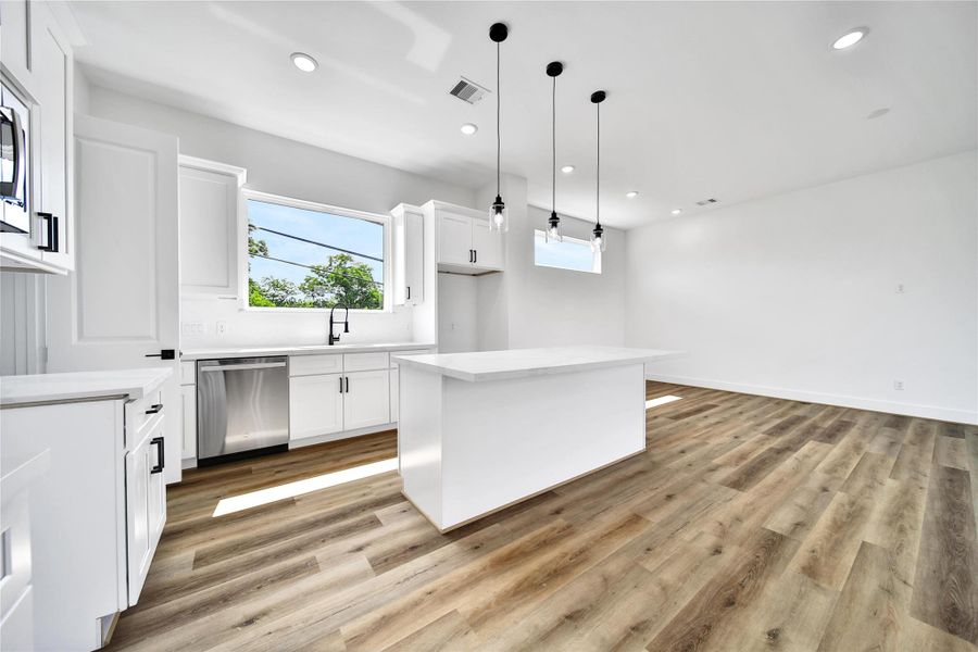 This kitchen features sleek white cabinetry, a central island, modern pendant lighting, and stainless steel appliances. It has ample natural light and a spacious open layout with elegant wood flooring.