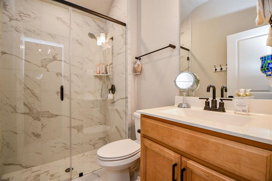 This bathroom features a modern design with a spacious glass-enclosed shower and elegant marble-patterned tiles. It includes a wooden vanity with a sleek countertop, black fixtures, and a well-placed mirror, offering a clean and stylish look.