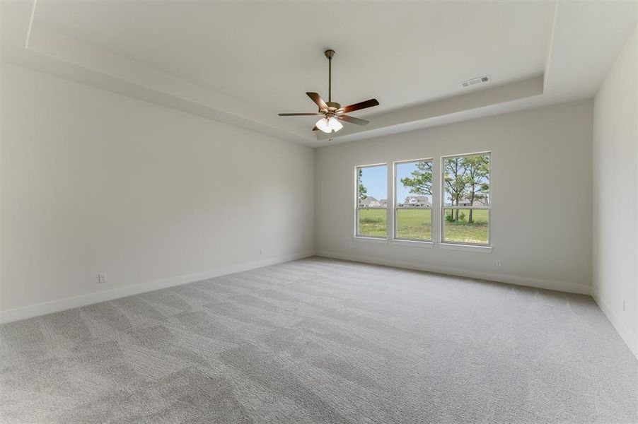 Spacious room featuring light gray carpeting, neutral wall finishes, a tray ceiling, an overhead fan with integrated lighting, and a three-panel window ensemble