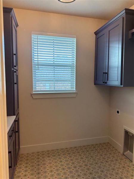 Utility space featuring patterned floor tile, built-in cabinetry with dark hardware, a window with blinds, and white baseboards