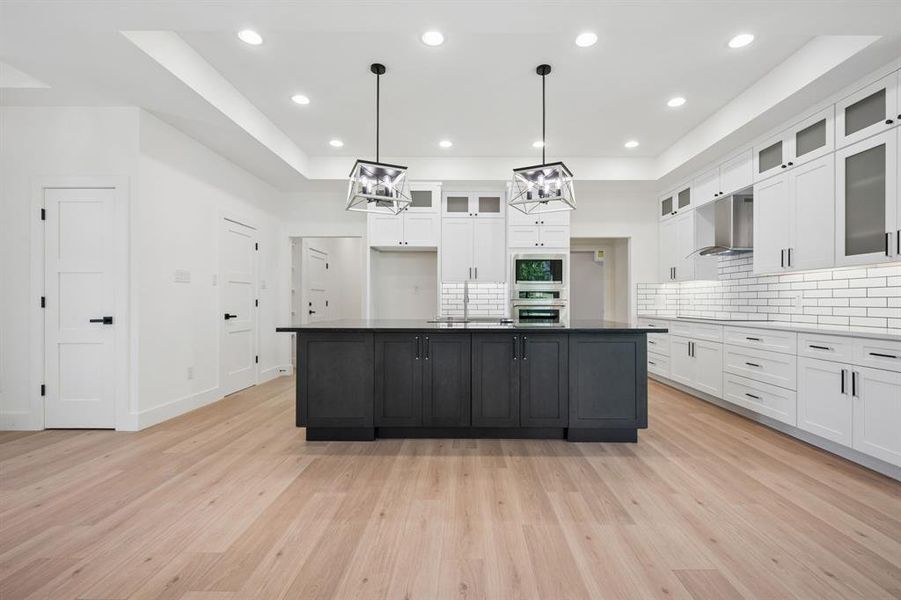 Two tone kitchen featuring backsplash, a kitchen island with sink, a raised ceiling, two tone color scheme, and glass fronted cabinets
