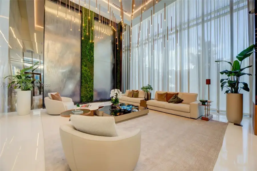 Double-height ceilings featuring floor-to-ceiling sheer drapery, a prominent indoor vertical living wall, a multi-panel decorative metal water feature, and linear pendant lighting fixtures
