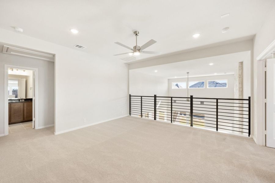 Open upstairs loft with carpet, ceiling fan, black metal railing, and view into bathroom Open upstairs loft with carpet, ceiling fan, black metal railing, and view into bathroom