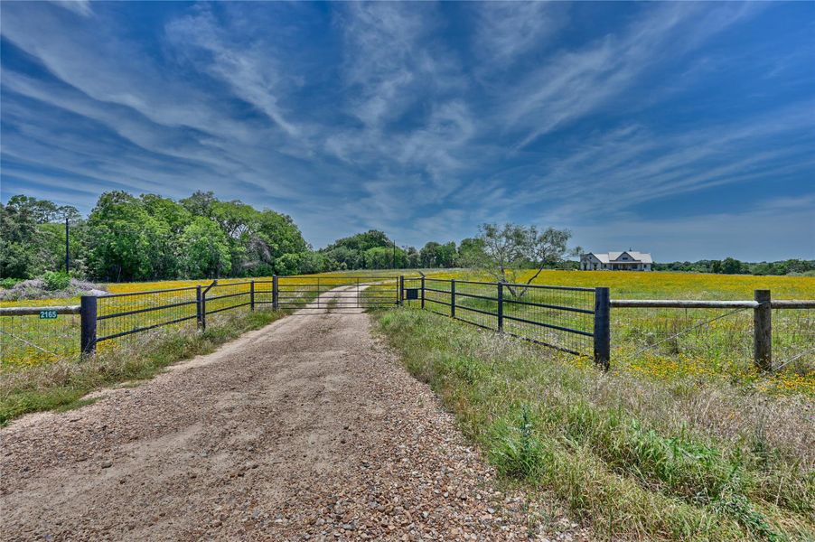 A private gravel driveway leads to a gated entry, opening to a sweeping view of a custom estate nestled amidst flowering fields.