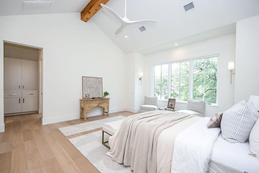 Bright and spacious bedroom with large windows, neutral decor, and light wood flooring. Features a cozy bed, seating area, and a stylish wooden console table. The room has a modern ceiling fan and a walk-in closet.