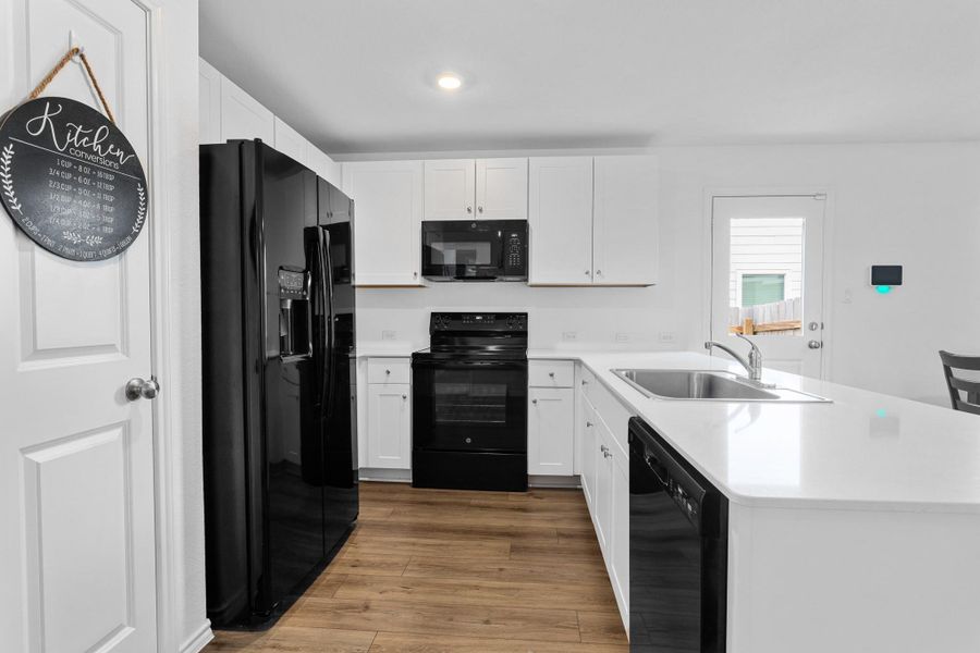 Modern kitchen featuring wood-finish flooring, white cabinetry, a built-in stainless steel sink, and black appliances including a refrigerator, range, microwave, and dishwasher
