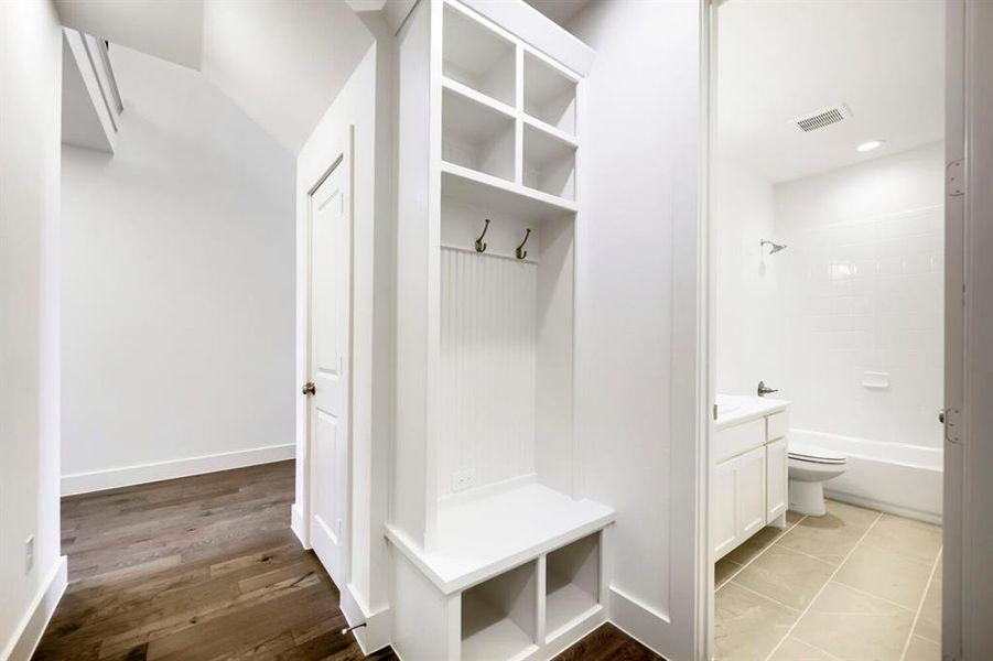 Hallway featuring wood-finish flooring, a built-in storage bench with coat hooks and cubbies, a paneled door, and a full bathroom with a tiled floor, white vanity, and tub with subway tile surround Hallway featuring wood-finish flooring, a built-in storage bench with coat hooks and cubbies, a paneled door, and a full bathroom with a tiled floor, white vanity, and tub with subway tile surround