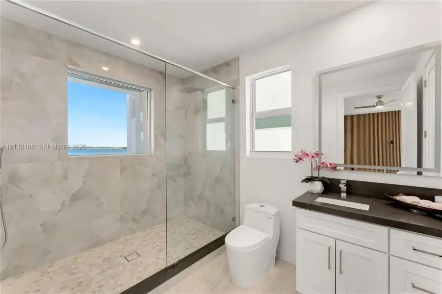 Bathroom featuring a walk-in shower with large-format wall tile and pebble-texture flooring, frameless glass enclosure, dual-pane window, and white cabinetry with a contrasting dark countertop Bathroom featuring a walk-in shower with large-format wall tile and pebble-texture flooring, frameless glass enclosure, dual-pane window, and white cabinetry with a contrasting dark countertop