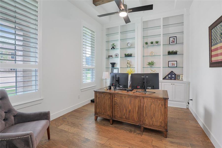Office space featuring wood-finish flooring, two large windows with plantation shutters, built-in shelving with glass inserts, integrated base cabinetry, and a ceiling fan