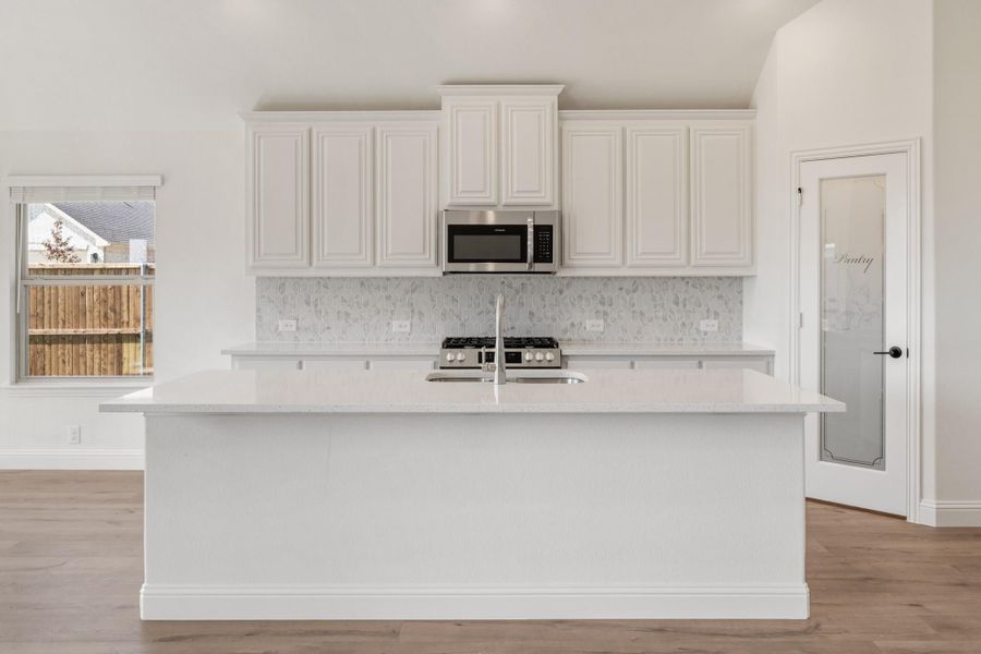 White kitchen with large island, raised-panel cabinets, tile backsplash, stainless microwave, and pantry door with glass panel. White kitchen with large island, raised-panel cabinets, tile backsplash, stainless microwave, and pantry door with glass panel.