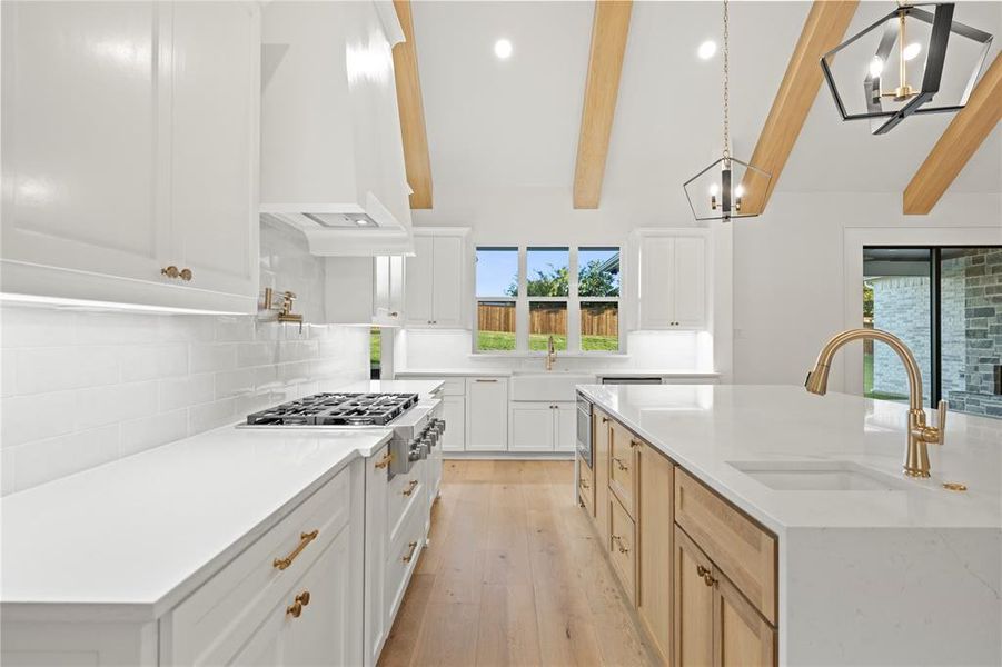 Spacious kitchen featuring vaulted ceilings with exposed wood beams, white cabinetry, a large kitchen island with integrated sink, stainless steel gas range, and wood-finish flooring