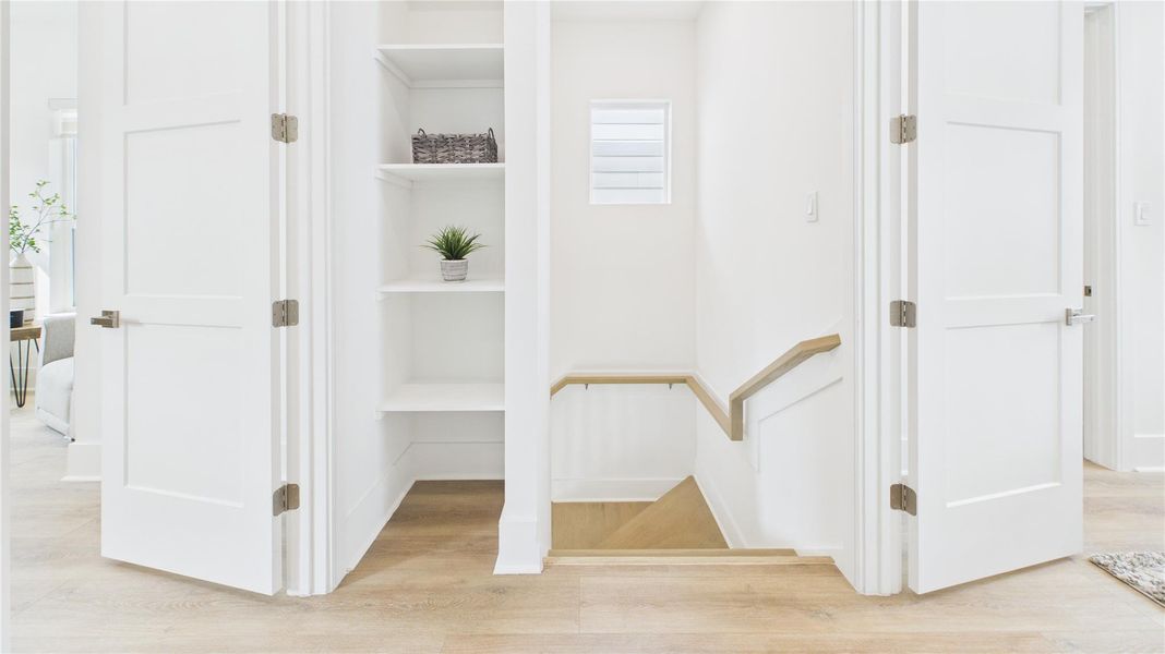 This photo shows a bright, clean hallway featuring light wood floors, white walls, and doors. There are open shelves with decorative items, and a staircase with a wooden railing leading down. The space is modern and inviting, with natural light from a small window.