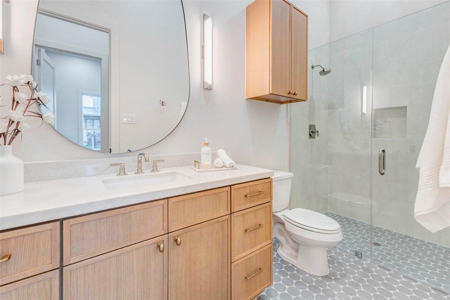 Elegant full bath with a frameless glass shower, light wood vanity, quartz countertop, and brushed nickel fixtures. The oversized oval mirror and geometric floor tile add designer flair to this sophisticated space.
