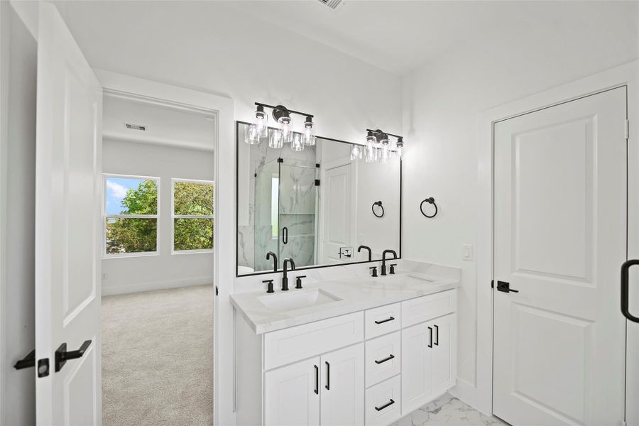 This bright, modern primary bathroom features a double vanity with sleek black fixtures, elegant lighting, and marble like quartz countertops. It opens to the spacious, carpeted primary bedroom with large windows offering a view of lush greenery.