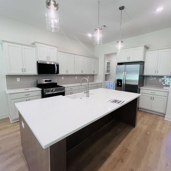 Kitchen featuring a large island with an integrated sink, white cabinetry, light countertops, wood-finish flooring, and stainless steel appliances