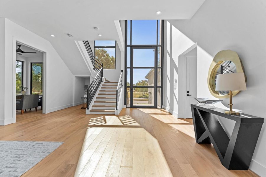 This photo showcases a modern, bright entryway with large windows, a sleek staircase, and light hardwood floors. The space includes a stylish console table with a lamp and decorative mirror, offering a welcoming and airy atmosphere.