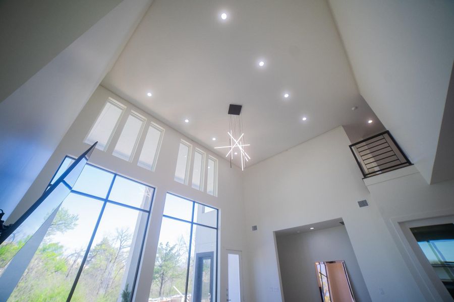 This home features a spacious, modern living area with a high ceiling and large windows, providing abundant natural light. A contemporary chandelier and a sleek balcony railing enhance the open and airy design.